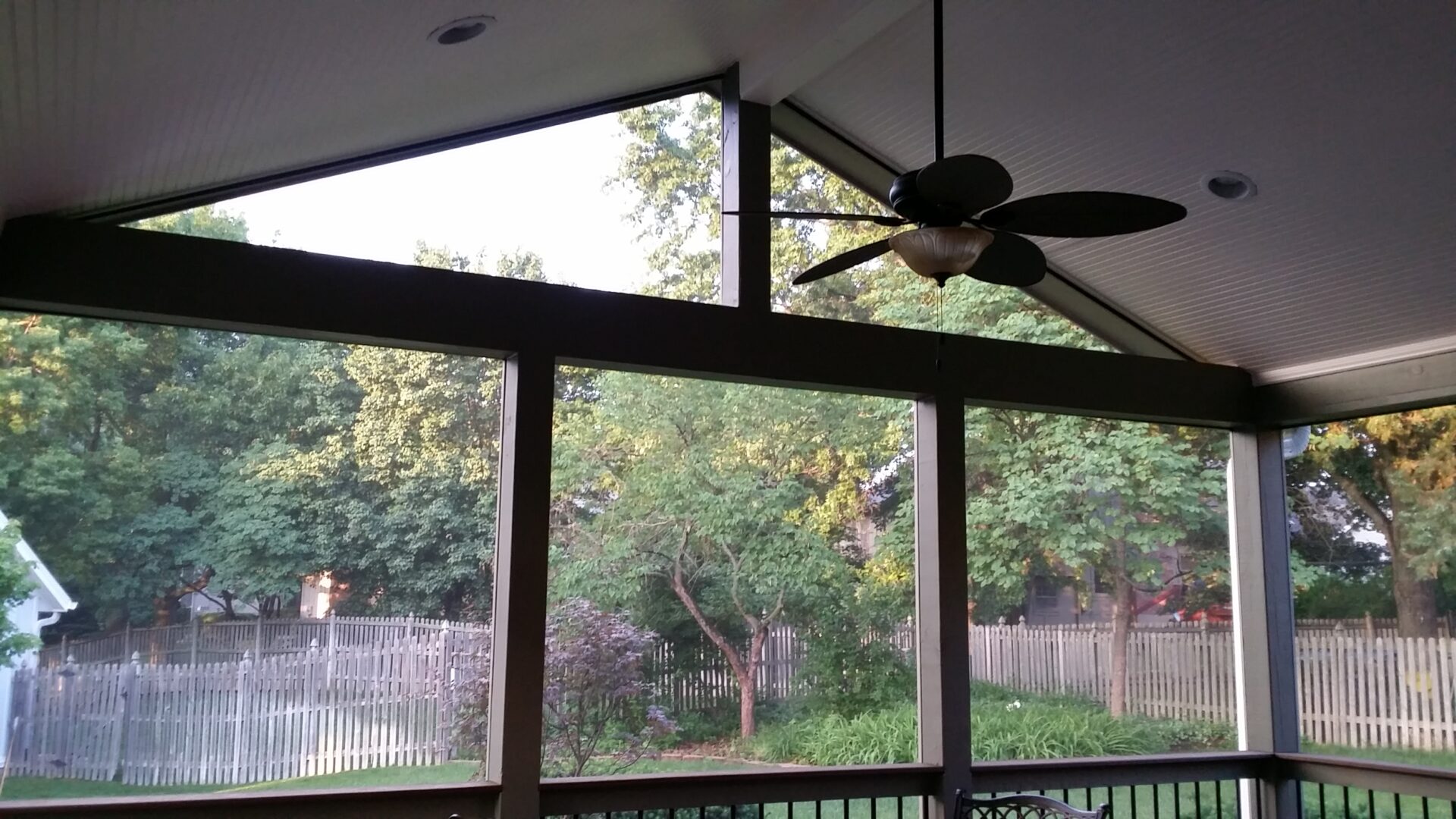 View of trees and a fenced yard through large screened windows and a ceiling fan.