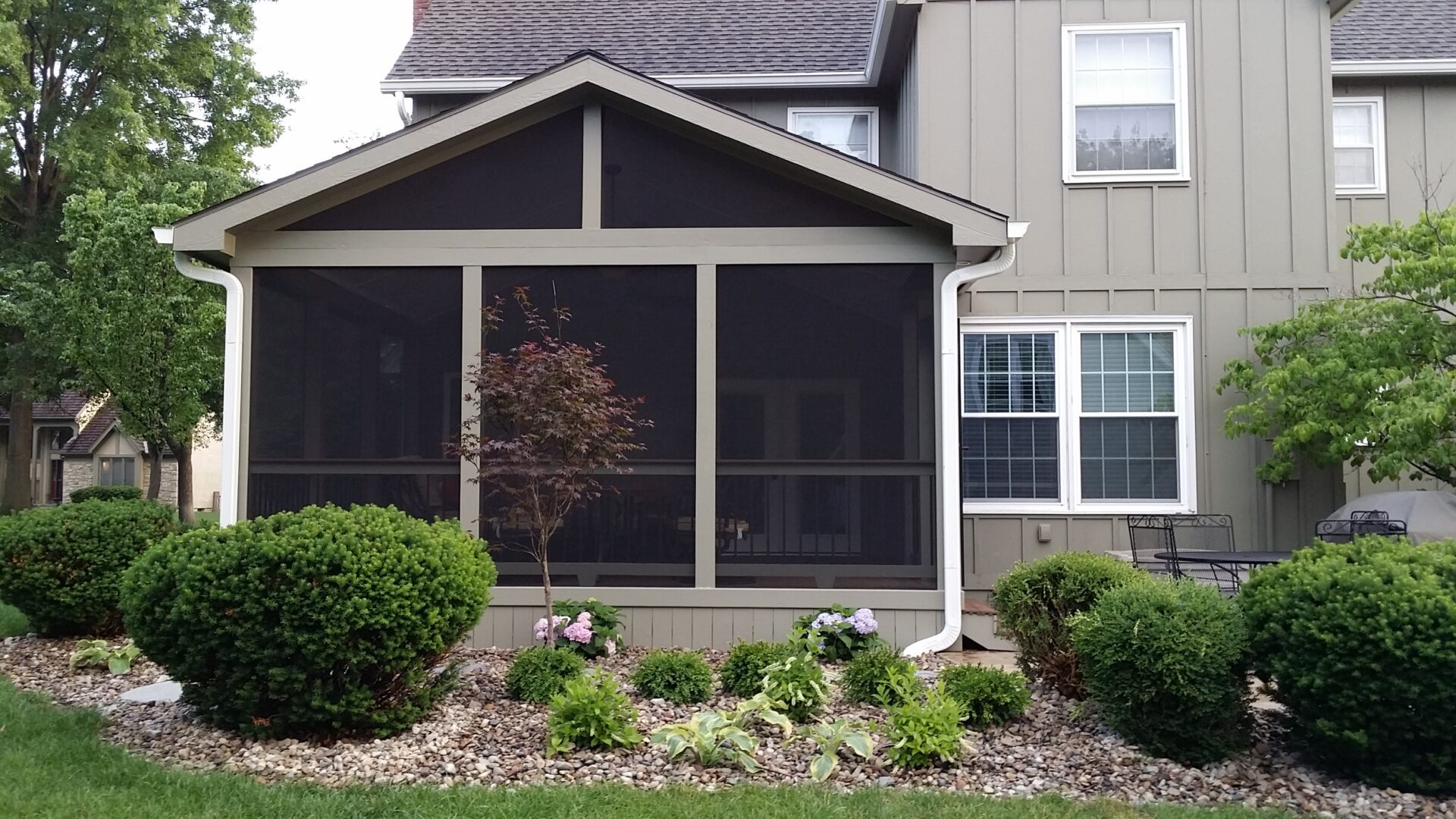 Screened porch with garden beds and shrubbery outside a house.