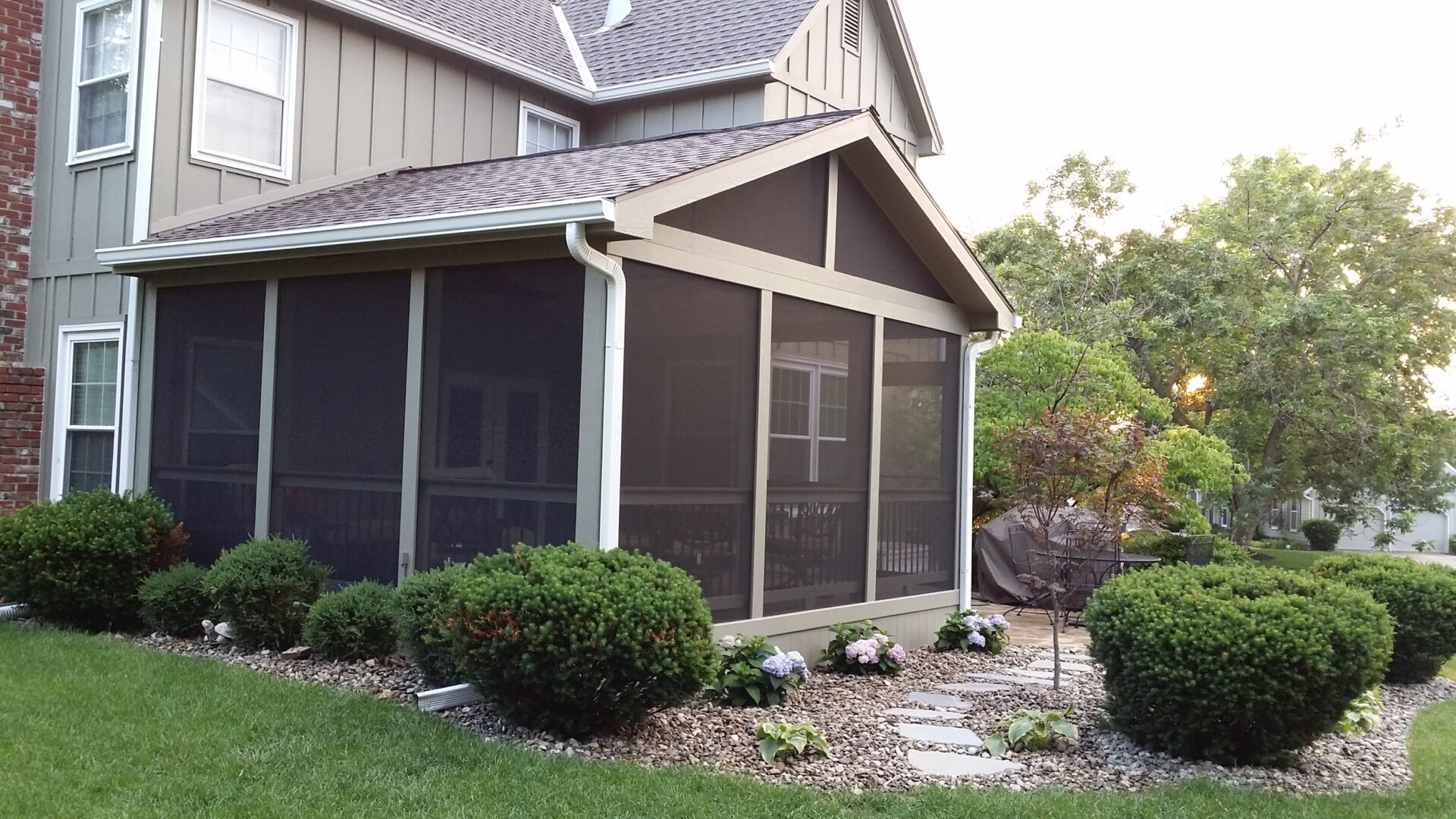 A screened porch attached to a house with surrounding landscaping.
