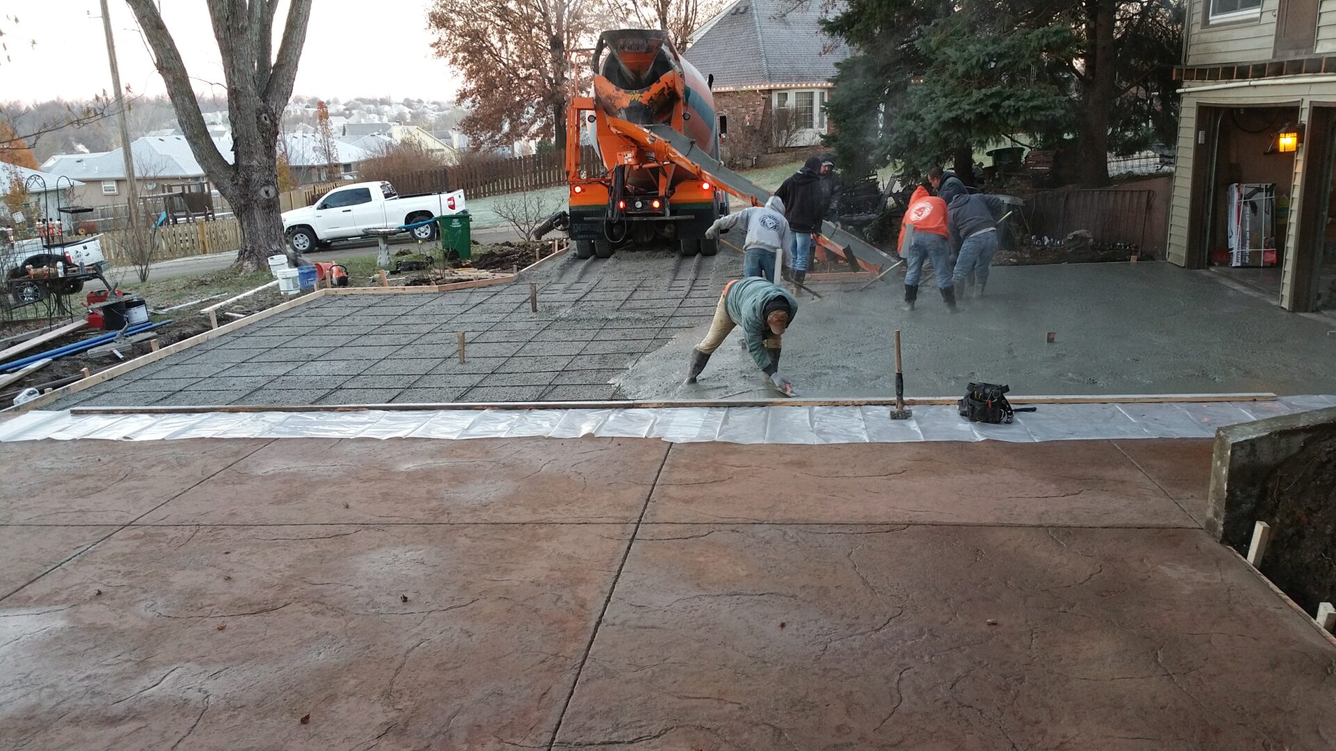 Workers smoothing freshly poured concrete on a driveway with an excavator in the background.