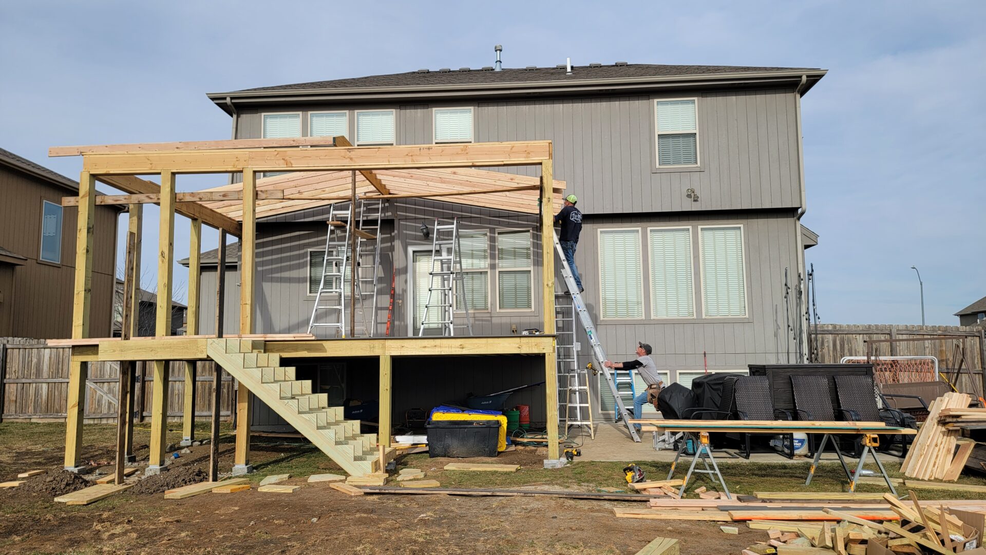 Two men constructing a wooden deck with ladders and tools.