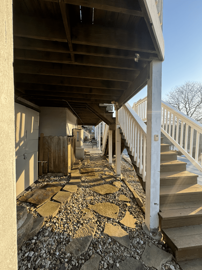 Sunlit stone pathway under an outdoor staircase beside a building.