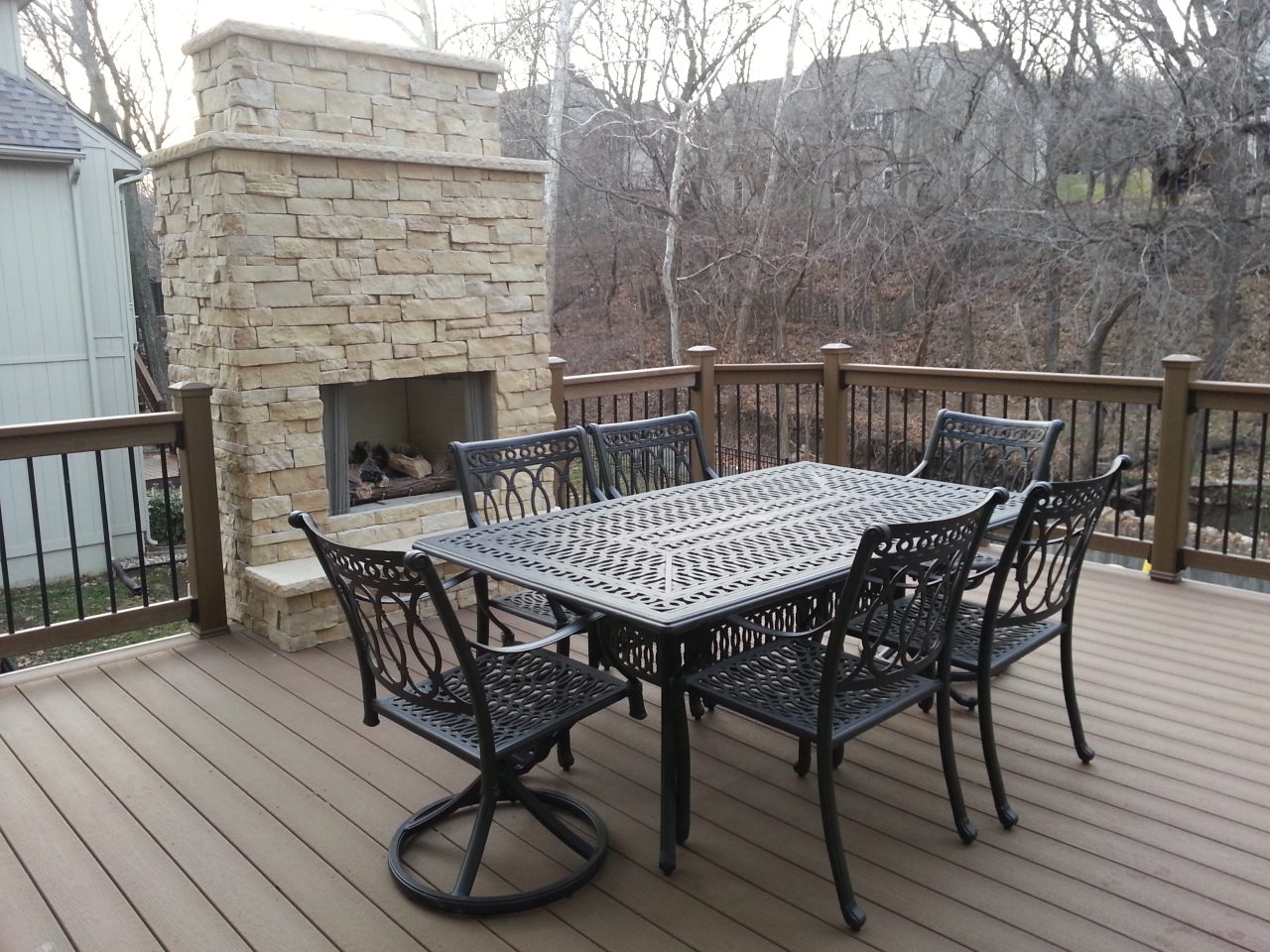 Outdoor dining table set with six chairs on a wooden deck with a stone fireplace.