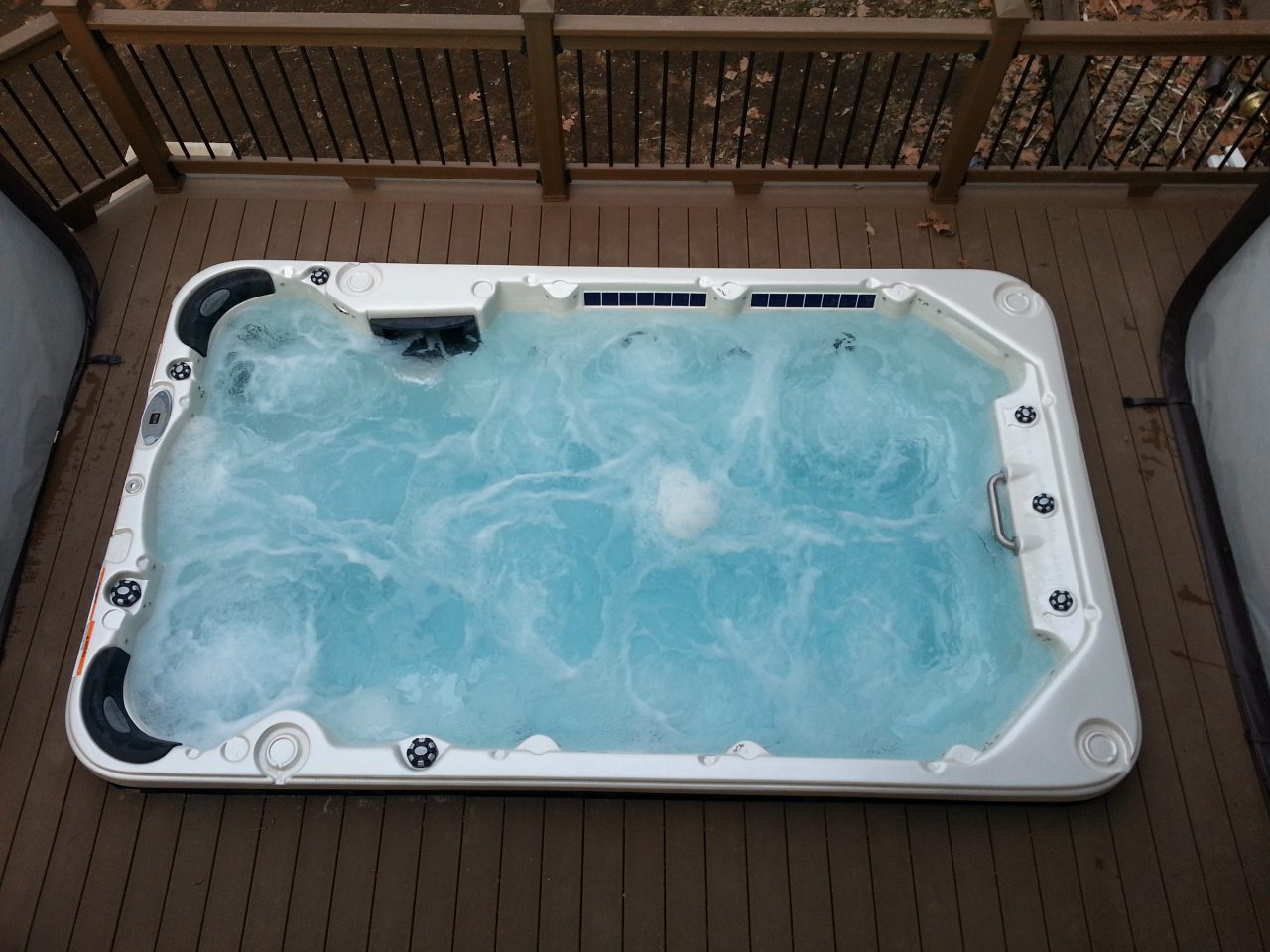 A bubbling rectangular hot tub on a wooden deck with a fence in the background.